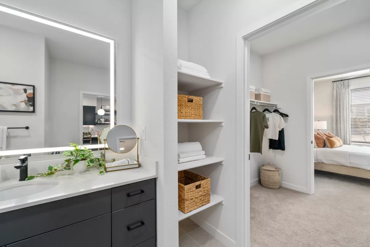 Bathroom vanity with backlit mirror and shelves stocked with toiletries and towels, with bedroom visible through the doorway.