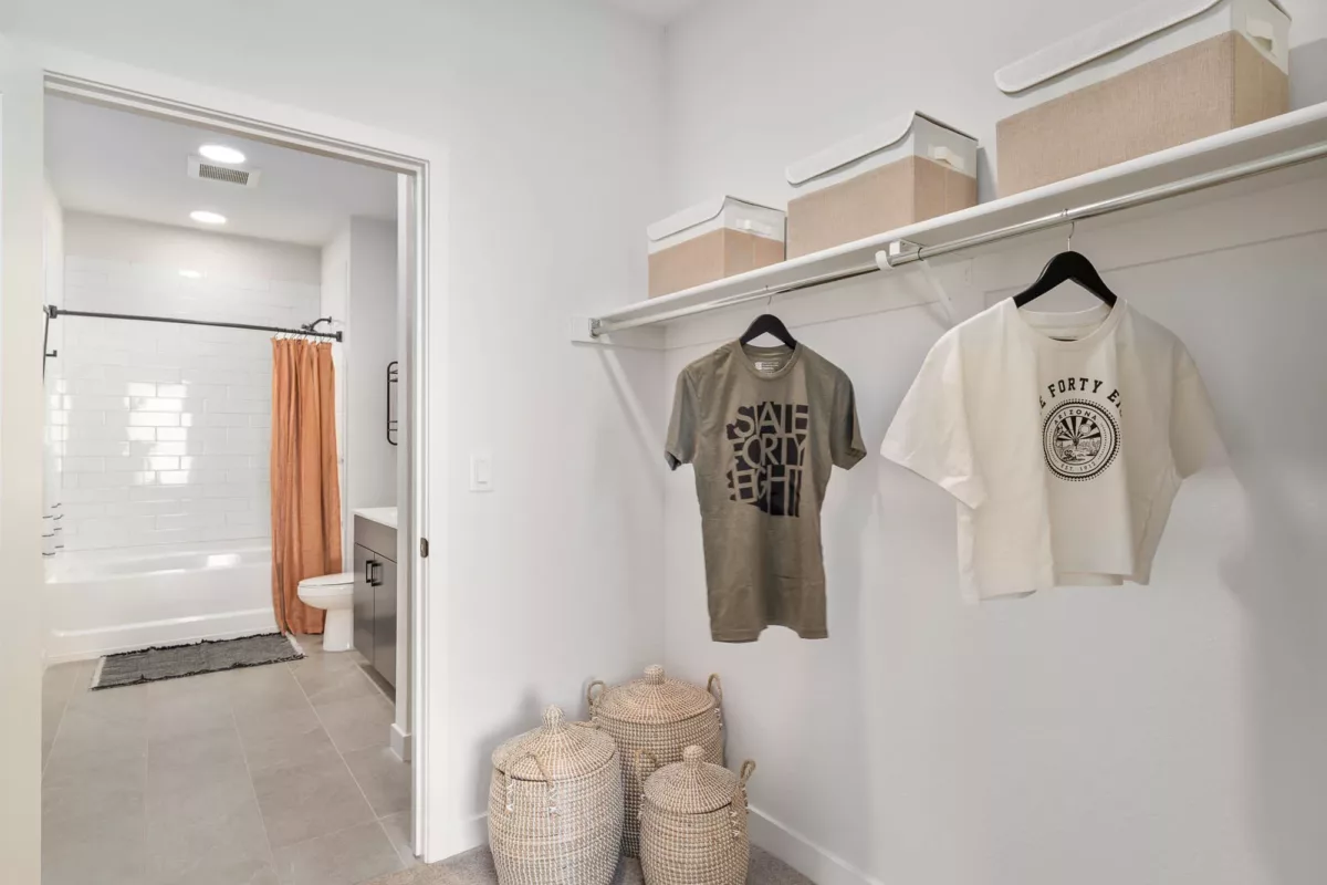 Walk-in closet with hanging clothes and baskets, leading into a bathroom with a tub and orange curtain.