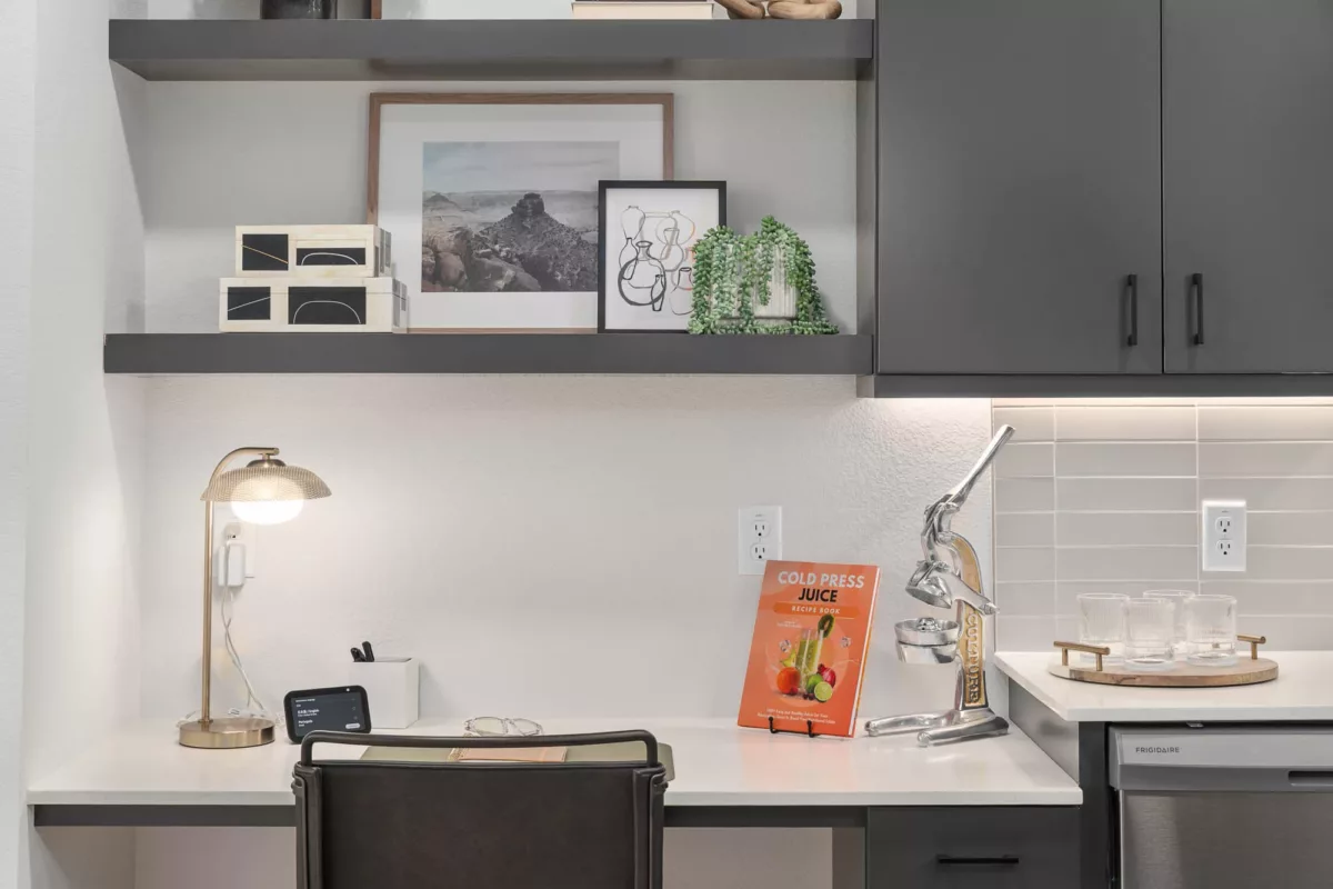 Compact kitchen workspace with desk, juicer, recipe book, and decorative shelving above.