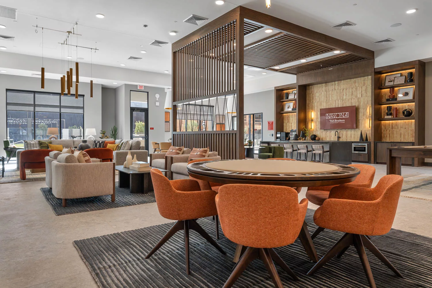 Contemporary clubroom with vibrant seating, poker table, and a wood-paneled partition, adjacent to an entertainment kitchen with bar and shelves.