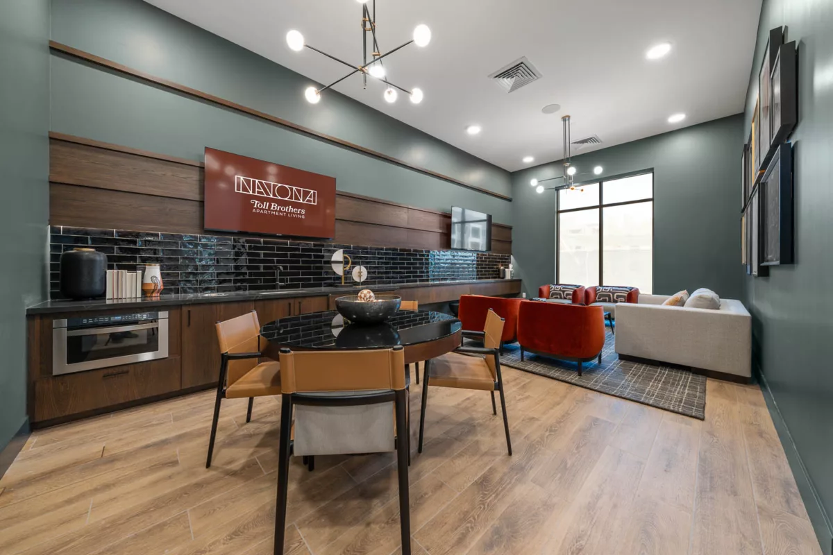 Sports lounge with a kitchenette, dining table, and red lounge chairs, set against dark green walls and decor.