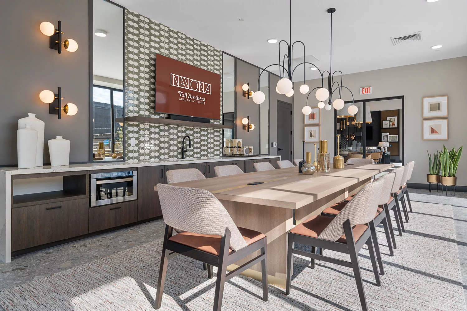 Dining room with a long wood table, upholstered chairs, decorative pendant lighting, and green tile backsplash near a kitchenette.